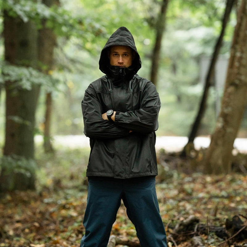 Man wearing Mil-Tec black waterproof rain jacket with hood in forest, hands crossed, outdoors on wet ground